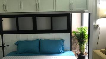 Uma cama com colcha listrada em branco e azul, cabeceira preta e duas almofadas azuis. Há uma planta em vaso num parapeito de janela à direita.