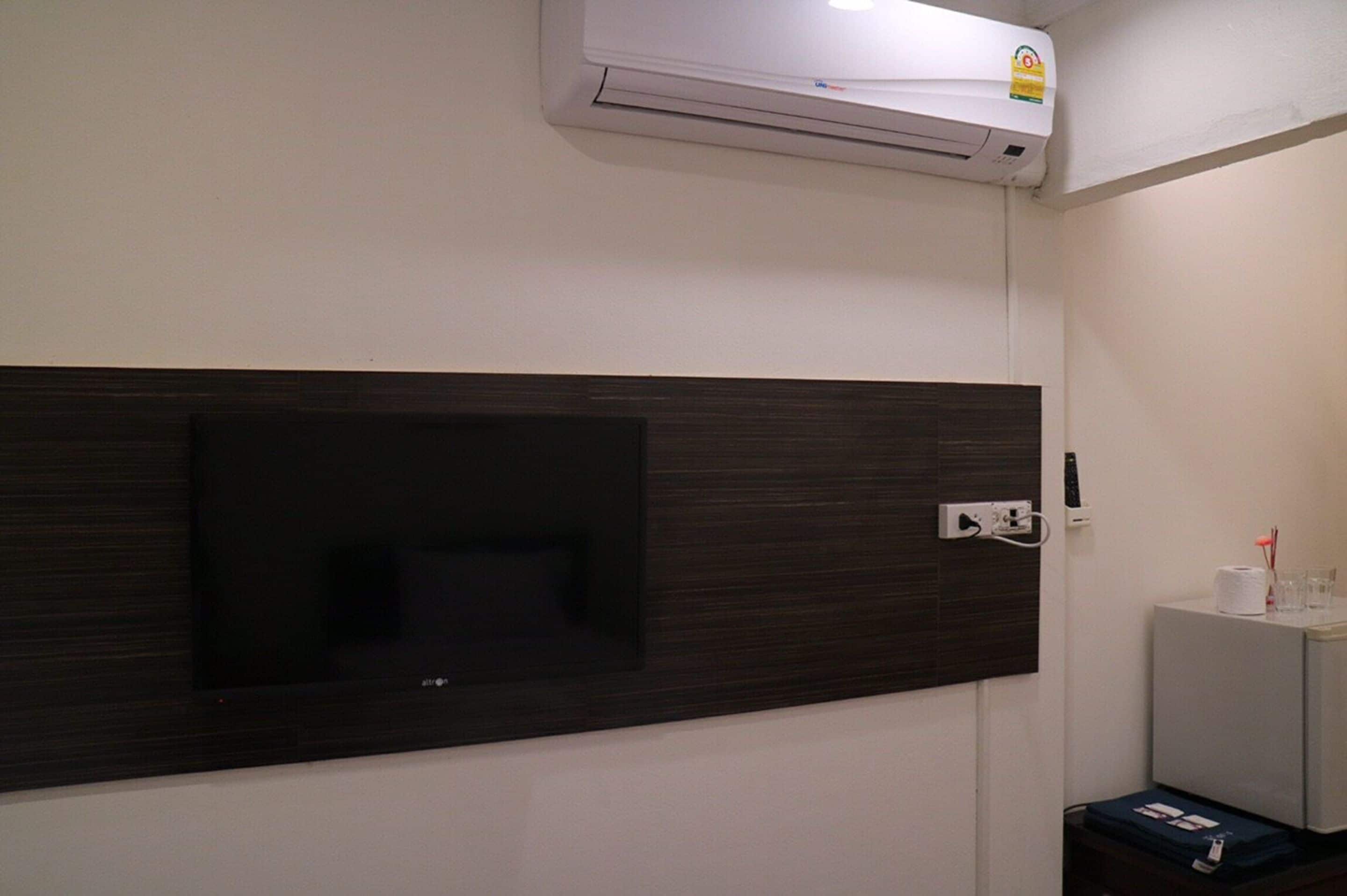 A room with a mounted flat-screen TV, an air conditioning unit, a power outlet, and a small cabinet with toiletries.
