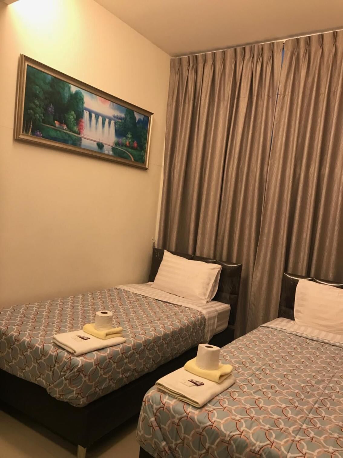 A hotel room with two beds, each with a patterned bedspread and pillows, a painting on the wall, and a large window with curtains.