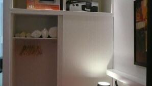 Quarto com uma cama, mesa de cabeceira com abajur, guarda-roupa com prateleiras e um quadro na parede.