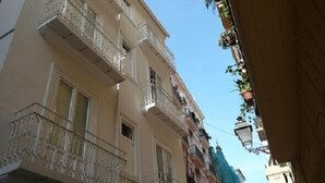 Uma rua estreita com edifícios altos, varandas com grades de metal e plantas penduradas.