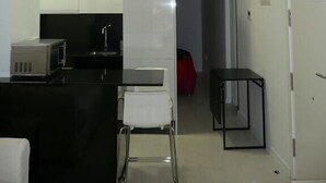 Cozinha compacta com armários brancos, bancada preta e uma cadeira branca. Há uma pequena mesa preta e uma porta à direita.