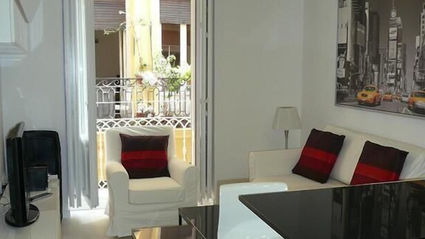 Uma sala de estar moderna com área de jantar, móveis brancos, varanda com plantas e uma pintura da paisagem urbana.