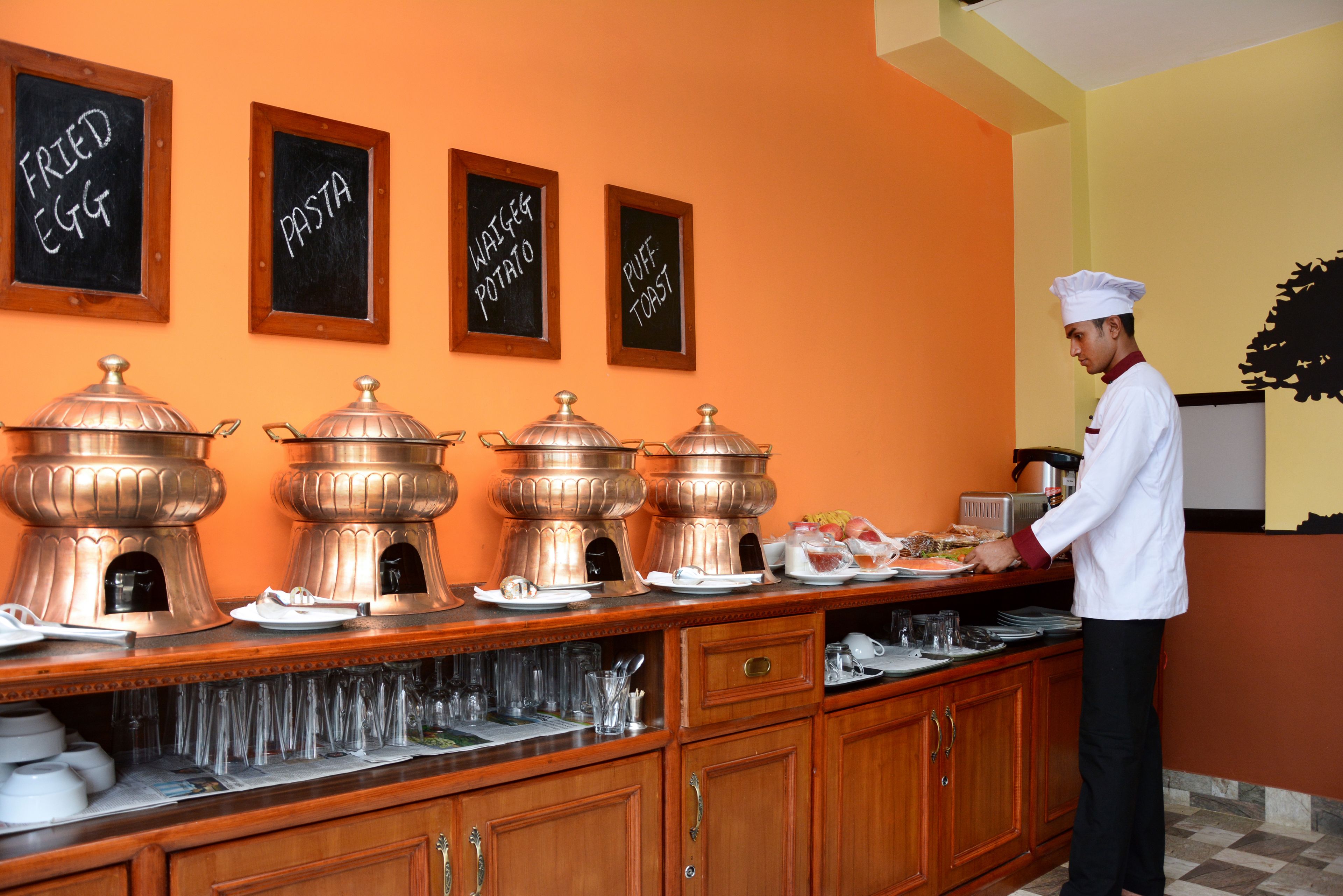 A buffet setup with labeled chafing dishes for fried egg, pasta, waffle potato, and puff toast.