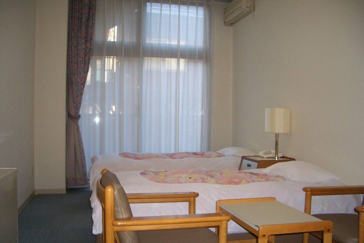 A hotel room with a bed, two wooden chairs, a small table, a bedside lamp, and a window with sheer curtains.