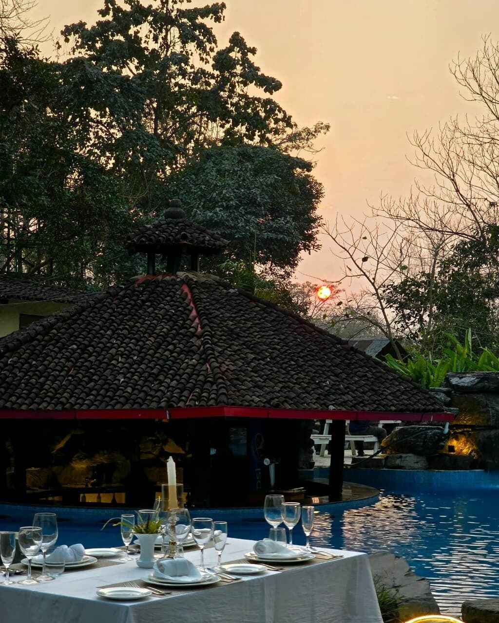 A dining area with a table set for a meal, a pool, and a building with a tiled roof.
