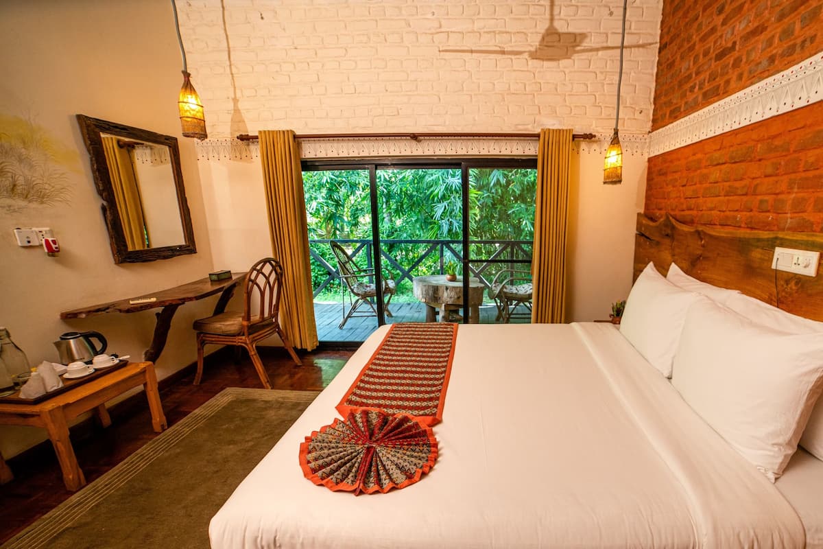 Une chambre d’hôtel avec un lit, une tête de lit en bois, une petite table avec une bouilloire, des chaises et une vue sur la verdure à travers une porte vitrée.