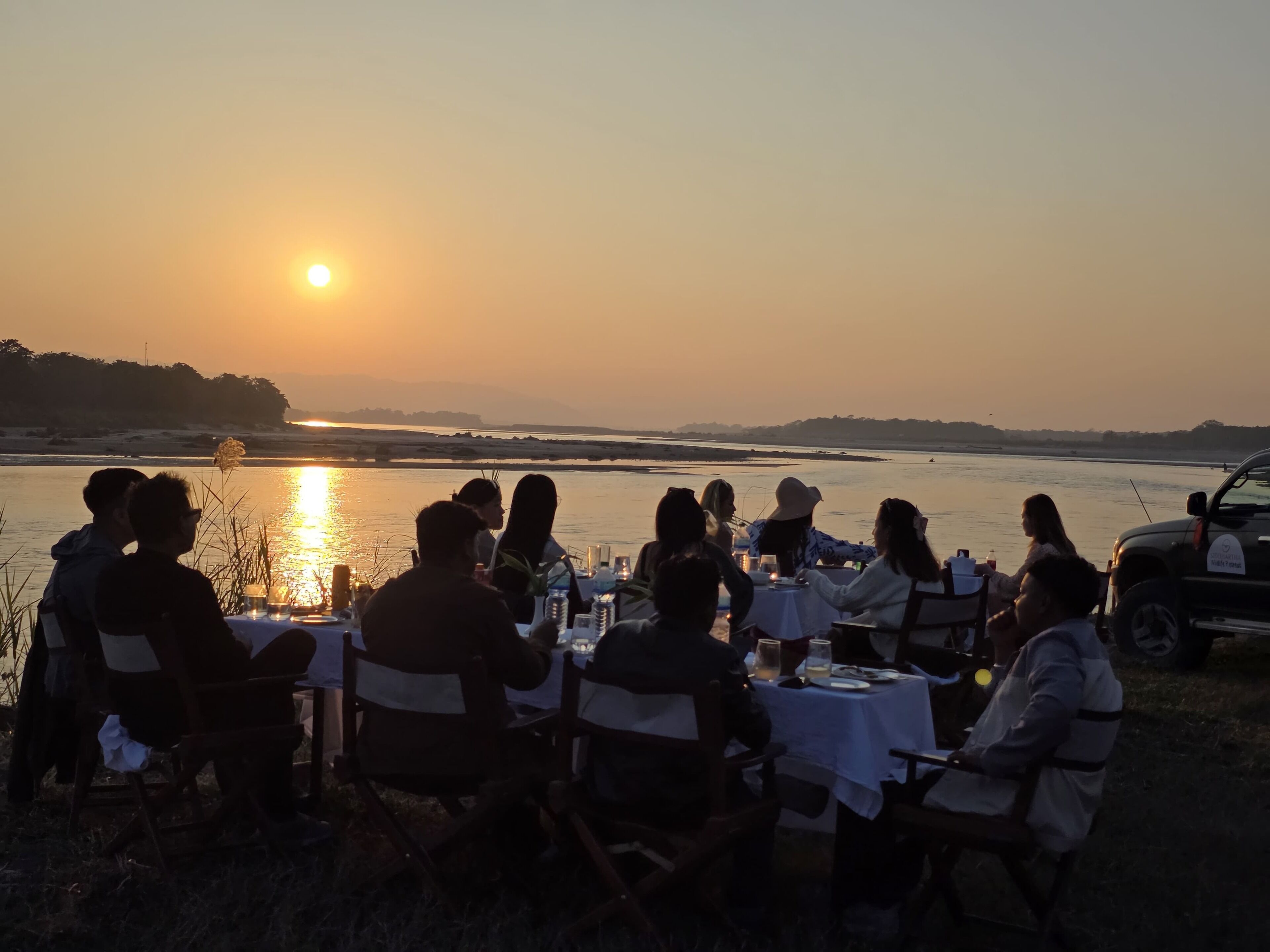 A group of people dining outdoors at sunset by a body of water, with tables set for a meal.