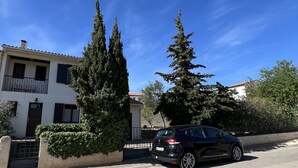 A residential street with a white building, a black car parked in front, and tall evergreen trees.