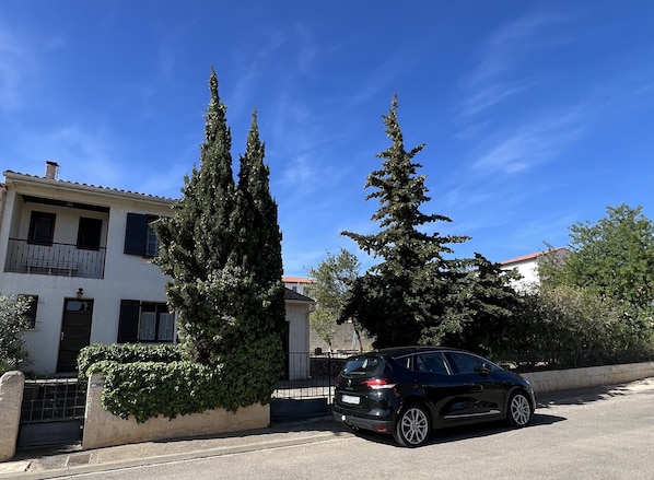 A residential street with a white building, a black car parked in front, and tall evergreen trees.