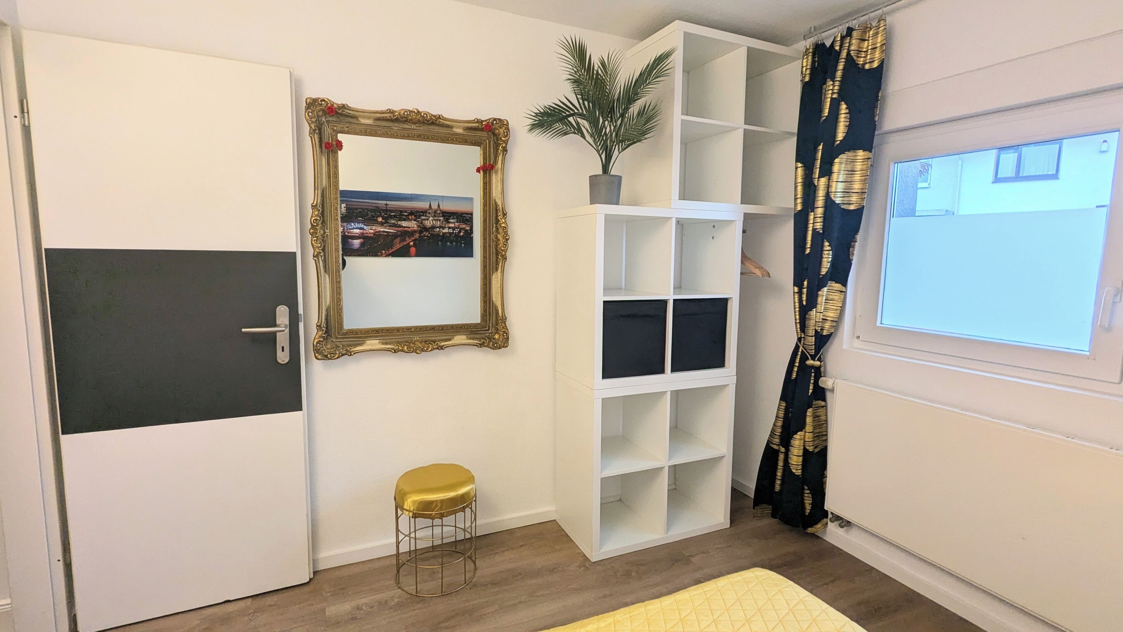 Une pièce avec une porte blanche, un miroir encadré d’or, un petit tabouret, une étagère blanche et une fenêtre ornée d’un rideau noir et doré.