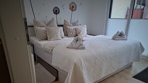 A neatly made bed with a white bedspread and pillows, a bedside table with a lamp, and a window with blinds. - Vacation Bungalow Bergblüte It is awarded with 4 **** (Bad Harzburg)