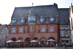 Il s'agit d'un bâtiment historique à la façade rouge, doté de nombreuses fenêtres et d'une tour centrale. On y trouve des tables et des chaises à l'extérieur, protégées par des parasols.