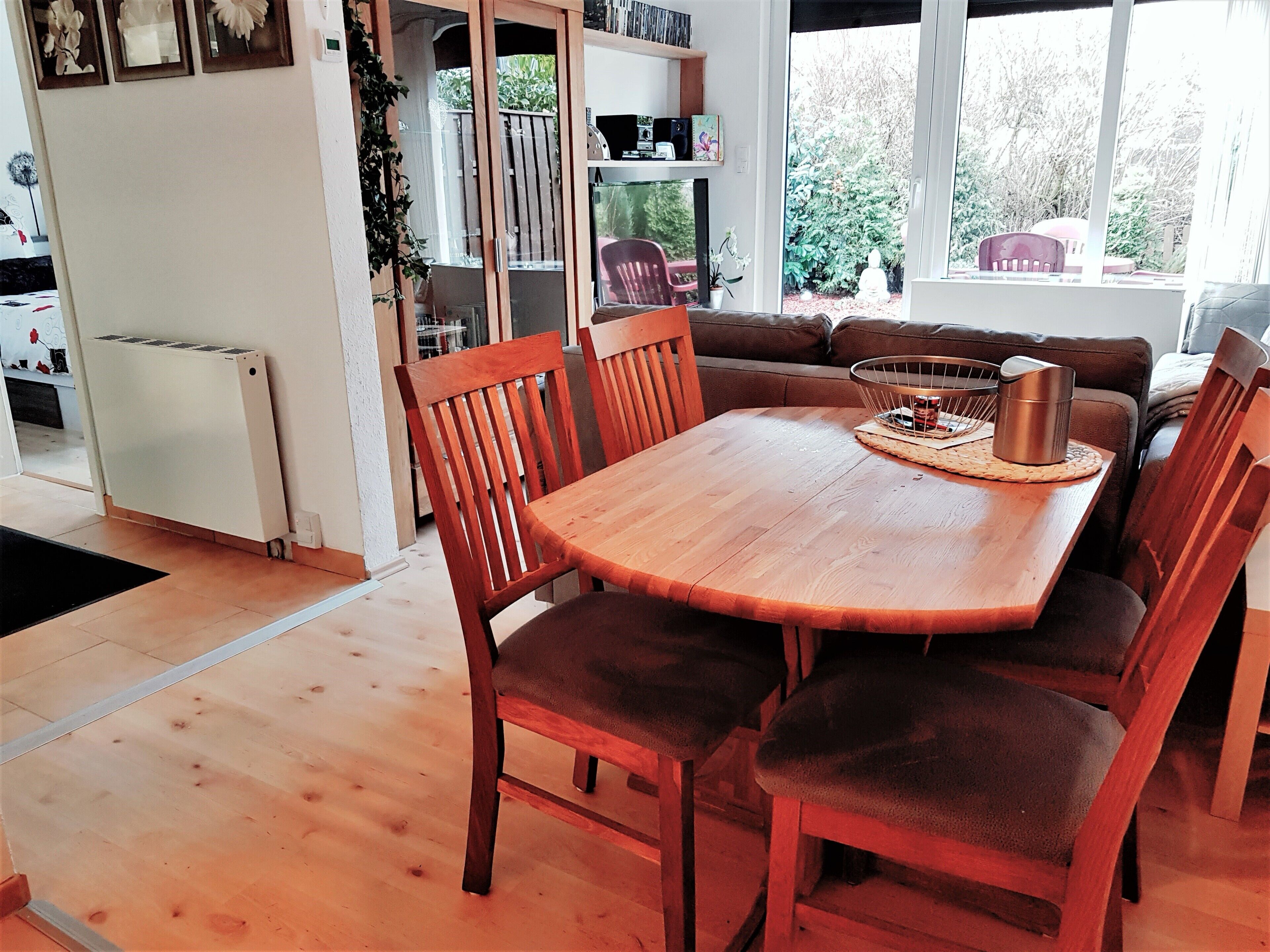 Un comedor con una mesa y sillas de madera, una pequeña cocina con refrigerador, y vista a un jardín a través de amplias ventanas.