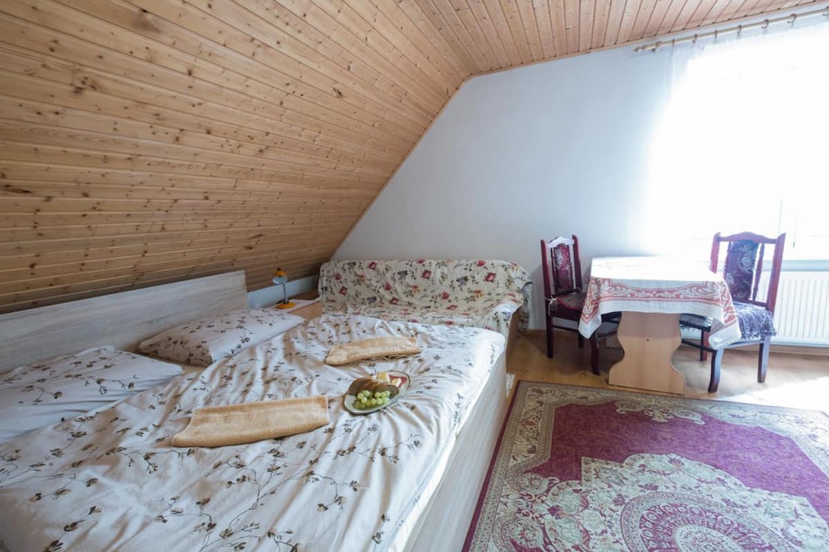 Une chambre avec un plafond en pente en bois, un lit avec une literie à motifs floraux, une table recouverte d’une nappe et une chaise.