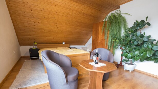 Ein gemütliches Schlafzimmer unter der Dachschräge, mit einem Bett, zwei grauen Sessel, einem Holztisch mit Flasche und Gläsern sowie einer Topfpflanze.