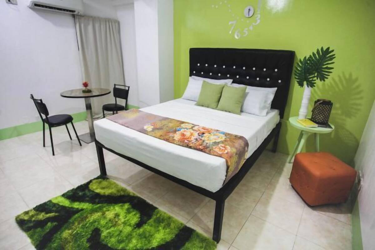 Ein Schlafzimmer mit einem Bett, einem runden Tisch mit Stühlen, einem kleinen Hocker und einer grünen Wand mit einer Uhr.