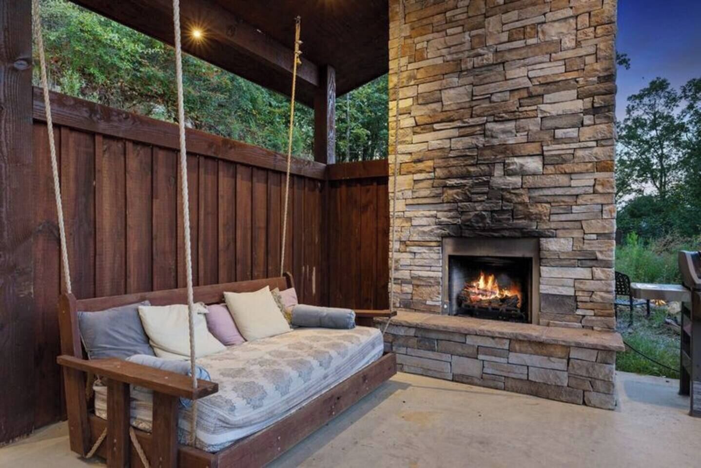A stone fireplace with a fire, a wooden swing seat, and a wooden deck.