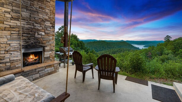 A stone fireplace, two Adirondack chairs, and a scenic view of a river and forest.