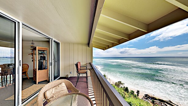 Un balcone con vista sull'oceano, una sedia in vimini, un tavolino e una porta scorrevole in vetro che dà su uno spazio interno con un armadio in legno e una poltrona.
