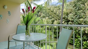 A round glass table with chairs, a vase with flowers, and a view of lush greenery and a palm tree.