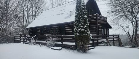 A log cabin with a steep roof, surrounded by snow-covered ground and trees.
