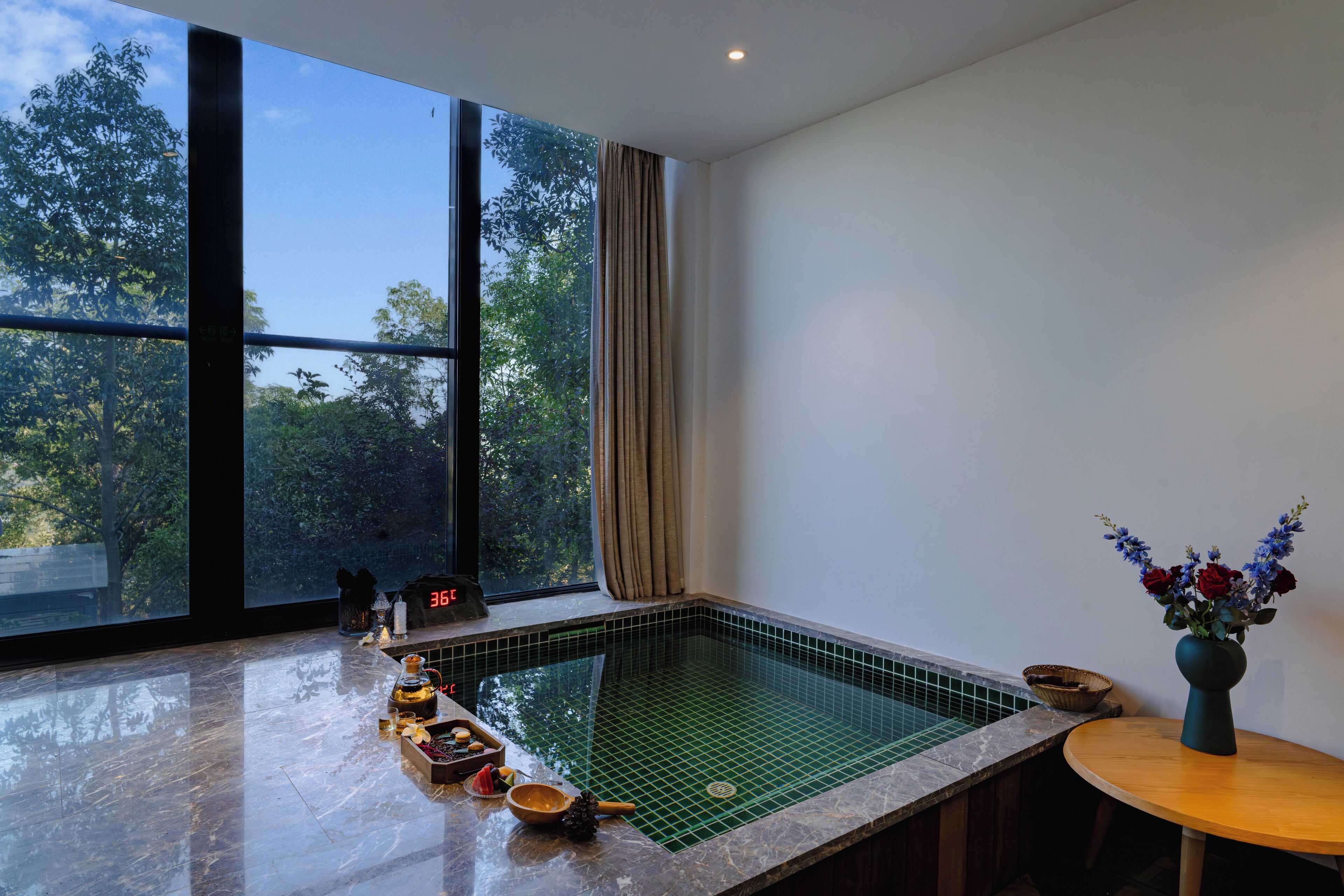 Une chambre moderne dotée d’une grande fenêtre, d’une petite piscine et d’une table en bois sur laquelle est posé un vase de fleurs.