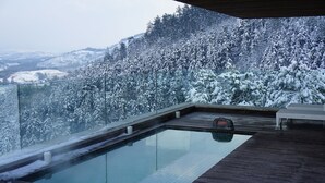 Une piscine à débordement moderne, dotée d’une terrasse en bois, entourée d’arbres couverts de neige et avec une chaîne de montagnes en toile de fond.
