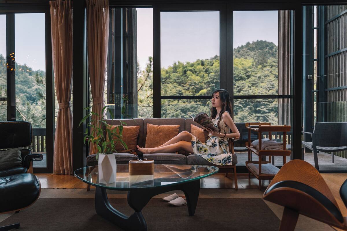 Ein modernes Wohnzimmer mit einem Glastisch, einer Couch und einem Sessel. Durch große Fenster ist ein Blick auf einen Wald zu sehen.