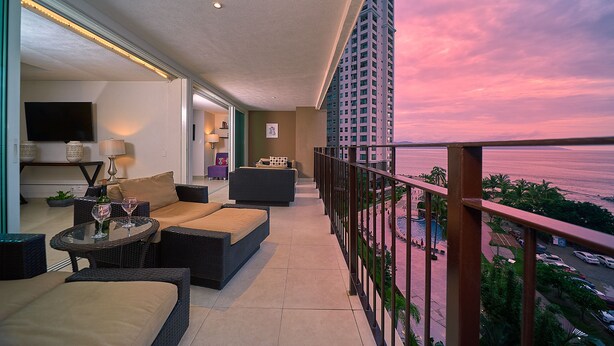 Un balcon offrant une vue sur l’océan au coucher du soleil, aménagé avec un canapé, des fauteuils et une petite table.