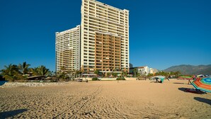 Un hôtel de grande hauteur doté de nombreux balcons, situé sur une plage bordée de palmiers et parsemée de parasols.