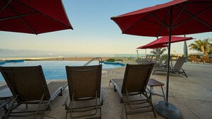 Un espace aménagé au bord de la piscine, avec des chaises longues et des parasols rouges, offrant une vue sur la plage et l’océan.