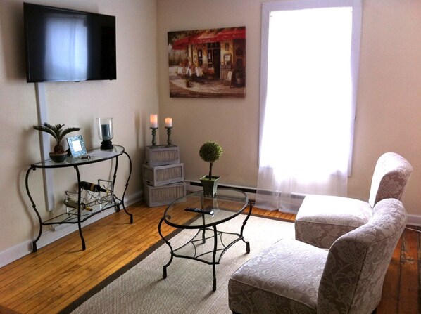 A living room with a TV mounted on the wall, a glass coffee table, a patterned armchair, a small table with candles, and a painting above the coffee table.
