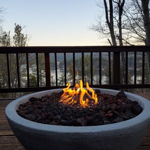 Un foyer en pierre avec des flammes, installé sur une terrasse en bois donnant sur un lac.