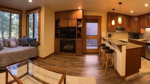A cozy living room with a sofa, a glass coffee table, and a fireplace. The kitchen features wooden cabinets, a refrigerator, and a bar area with stools.