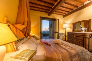 A hotel room with a bed, a bedside table with a lamp, a wooden headboard, a window with curtains, and a view of the outside.