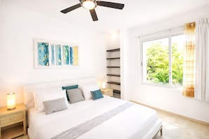 Une chambre à coucher comprenant un lit, des tables de chevet, un ventilateur de plafond, une fenêtre avec des rideaux et une œuvre d’art abstraite accrochée au mur.