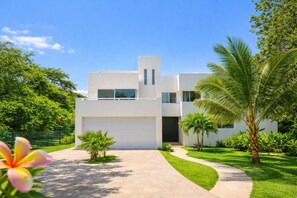 Une maison moderne à deux étages, avec une façade blanche, un garage et une pelouse bien entretenue agrémentée de palmiers.