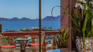 A balcony with wicker chairs and a table, overlooking a coastal view with mountains and the sea.