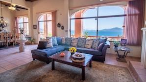 A spacious living room with a large sofa, a coffee table, and a view of the sea.