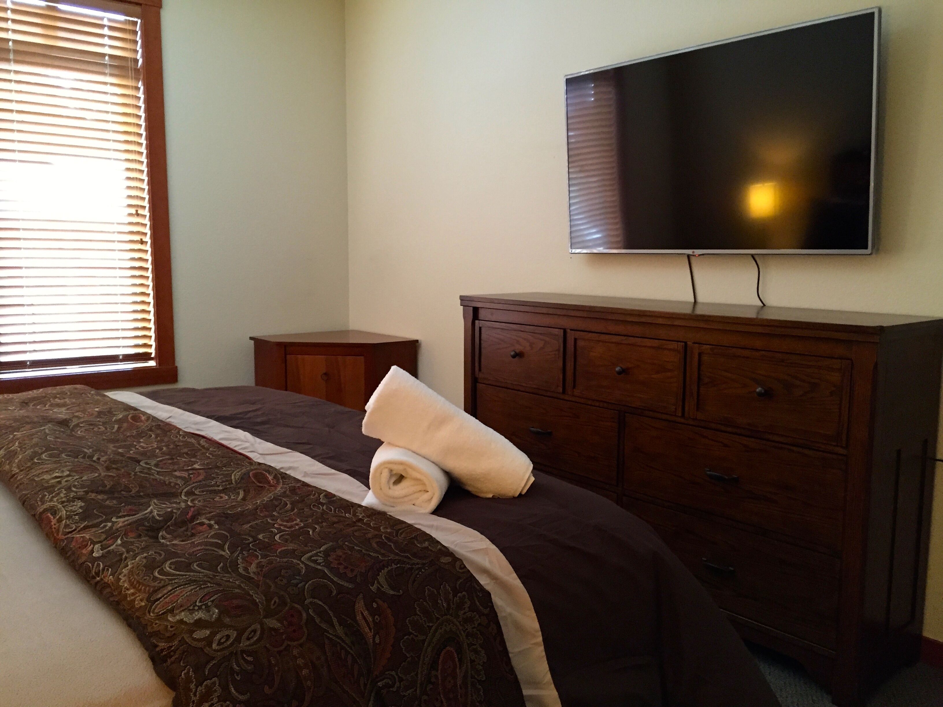 A bedroom with a bed, a wooden dresser, a television mounted on the wall, and a window with blinds.