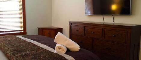 A bedroom with a bed, a wooden dresser, a television mounted on the wall, and a window with blinds.
