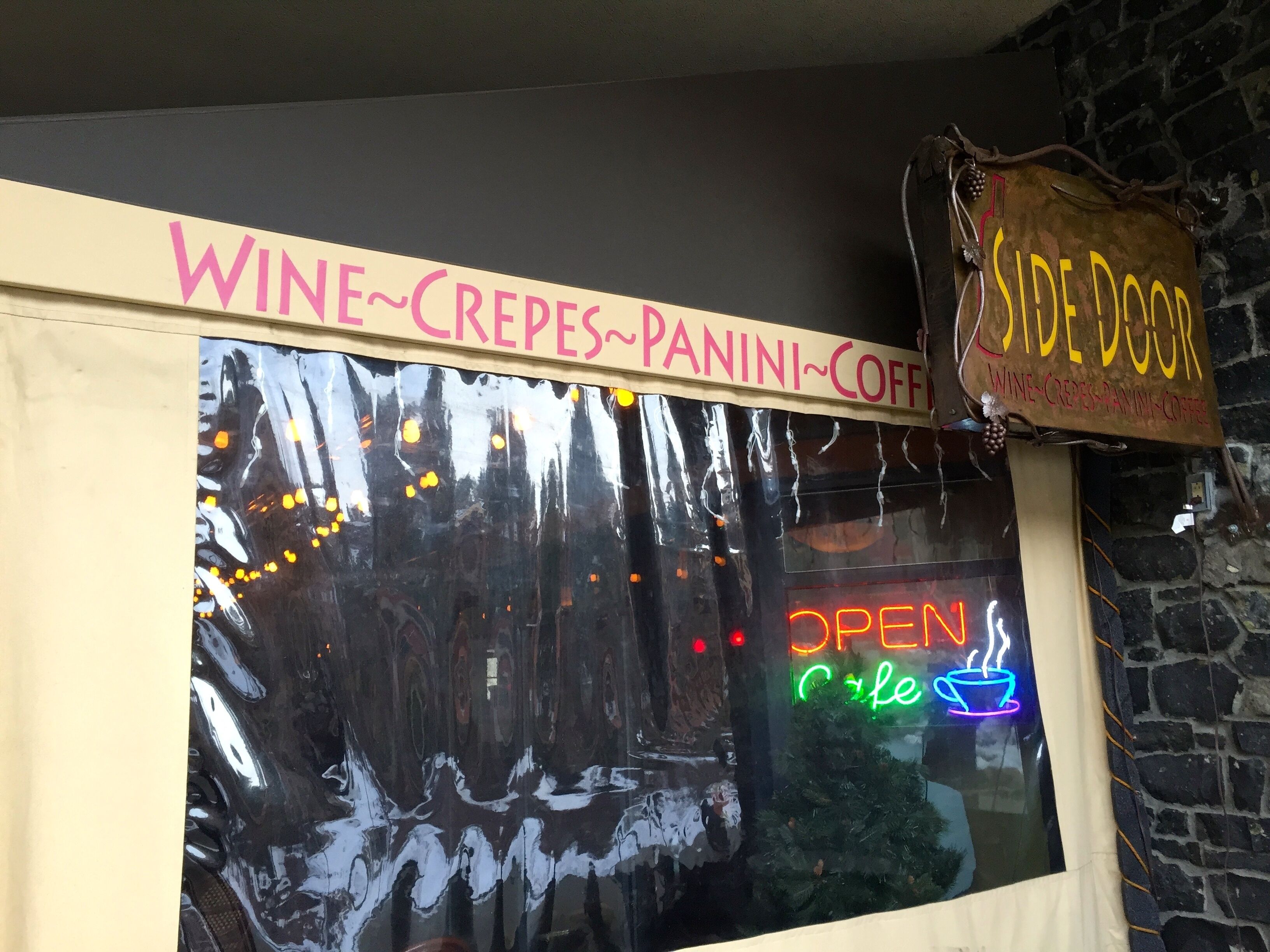 Ein Geschäft mit einer Schaufensterreklame mit der Aufschrift „WINE-CRÈPES-PANINI-COFFEE“ und einem leuchtenden Neonzeichen, das anzeigt, dass die Einrichtung geöffnet ist.