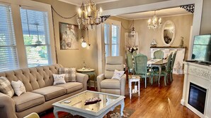 A living room with a sofa, armchairs, a coffee table, and a fireplace. There is a dining area with a table and chairs. - Stylish Coastal Escape | 3BR Shabby Chic + Optional Cottage, Walk to Beach & Fun (Gulfport)