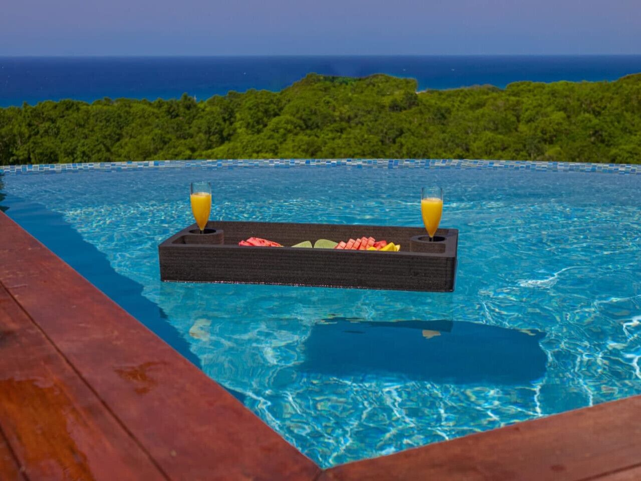Ein Pool mit einer schwimmenden Bar und Getränken, umgeben von einer Holzterrasse und Blick auf das Meer und die grüne Landschaft.