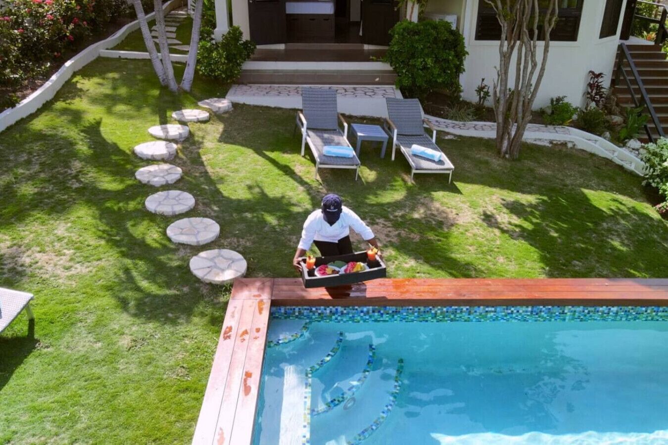 Un área junto a la piscina con un camarero sirviendo bebidas, una piscina con un borde de mosaicos y un césped bien cuidado con piedras para pisar.