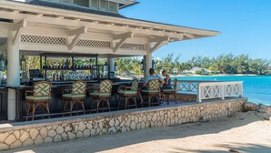 Un bar en la playa con sillas de mimbre y un muro de piedra, con vistas a un mar azul cristalino y palmeras.