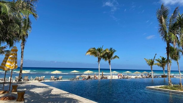 Un área junto a la piscina con sillas de descanso y sombrillas, palmeras y vista al océano.