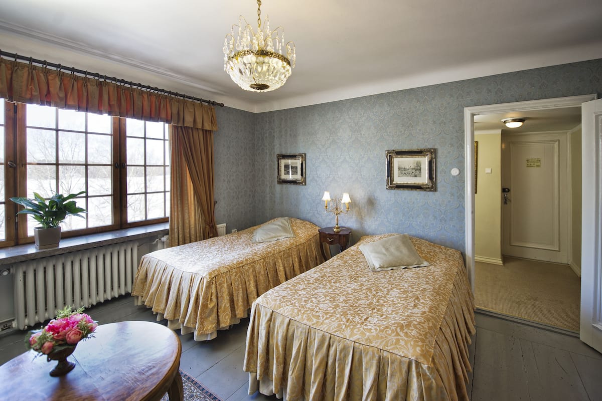 Een hotelkamer met twee bedden, een kroonluchter, een radiator en een raam met gordijnen.