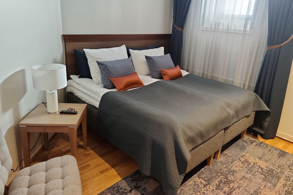Een verzorgde slaapkamer met een bed, nachtkastje, lamp en uitzicht op het groen door dunne gordijnen.
